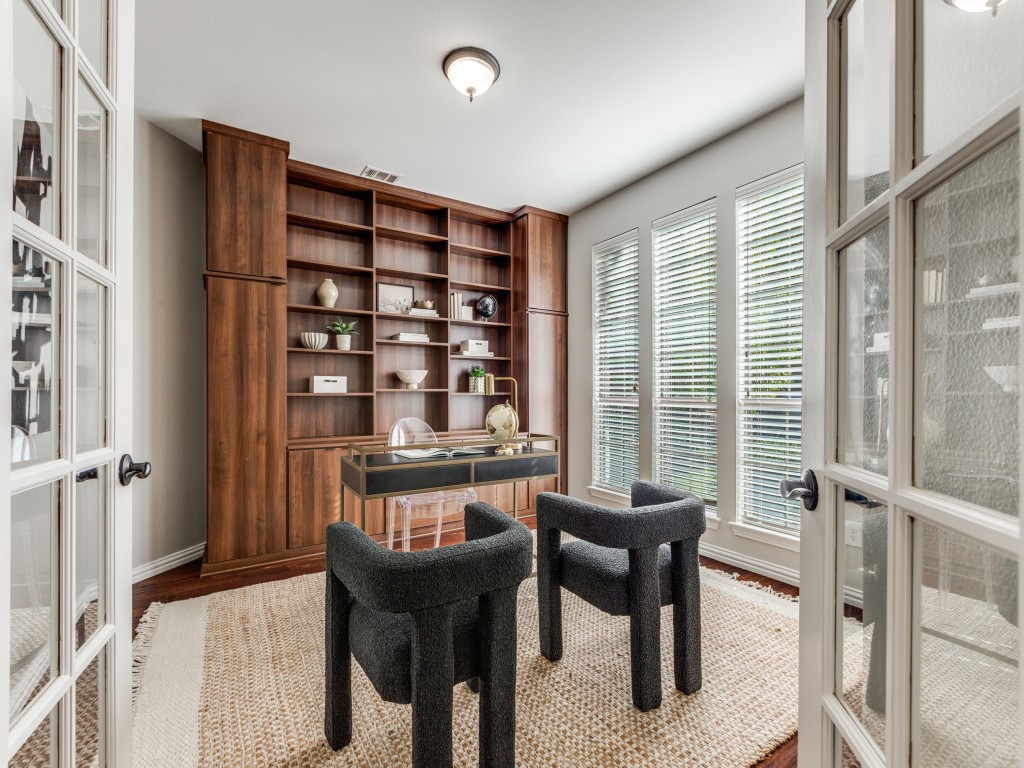 Modern home office with a wooden bookshelf, glass desk, two black chairs, a beige rug, and large windows with blinds letting in natural light.