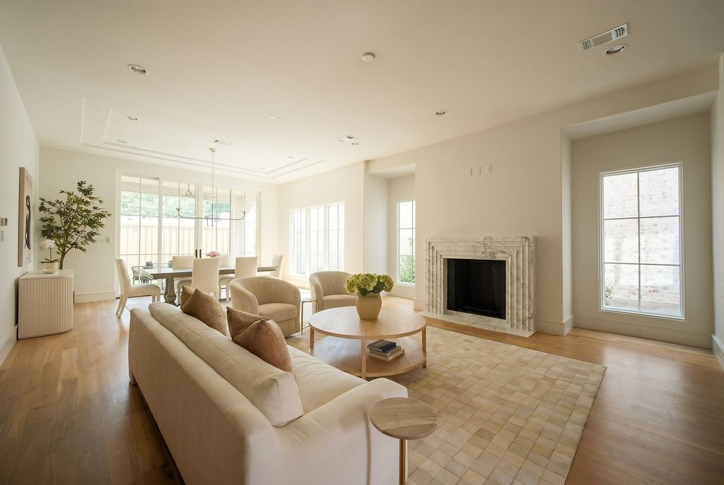 Spacious living room with cream-colored sofa, round coffee table, beige rug, dining area, marble fireplace, wooden floor, and large windows letting in natural light.