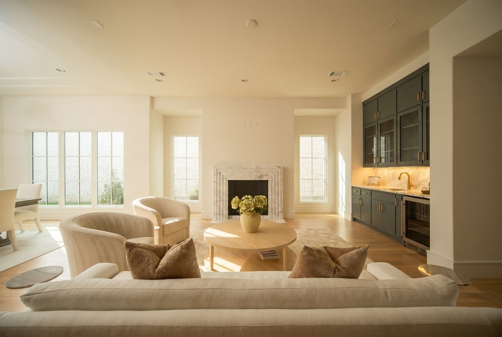A bright living room with large windows, a white sofa, two armchairs, a round coffee table with flowers, a fireplace, and a kitchen area with dark cabinets.