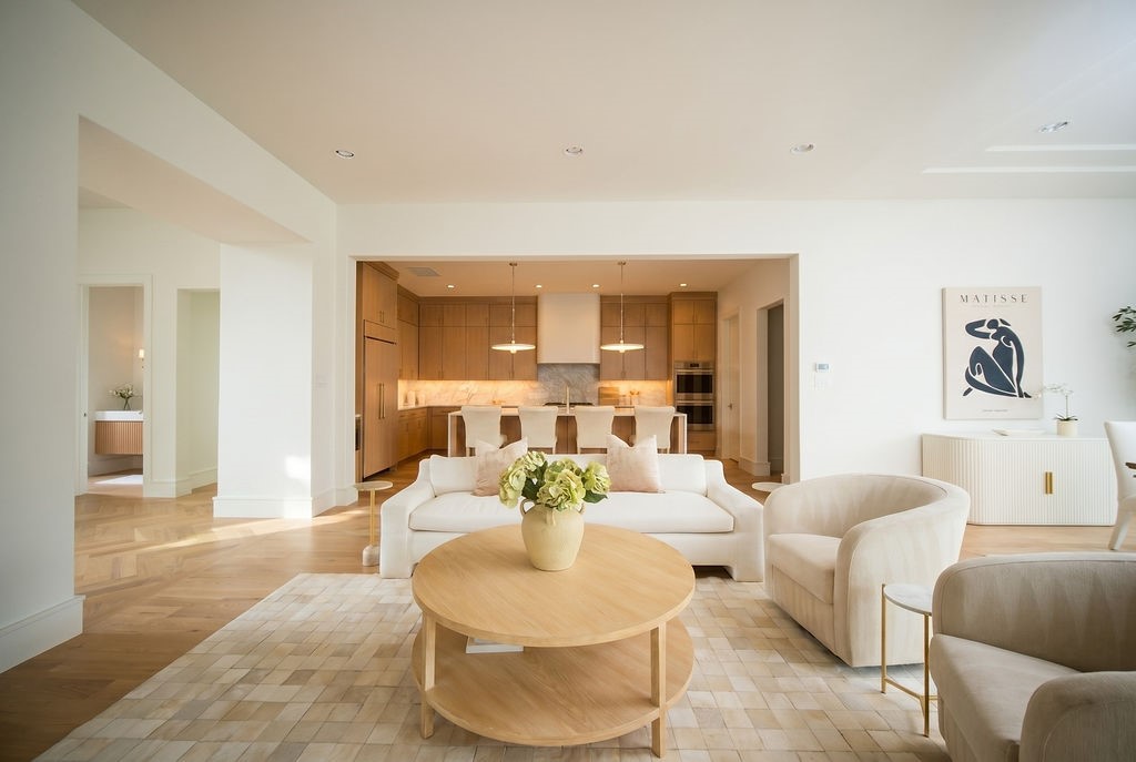 Bright modern living room with neutral furniture, a round coffee table, and an open kitchen in the background; wall art and a potted plant add decorative touches.