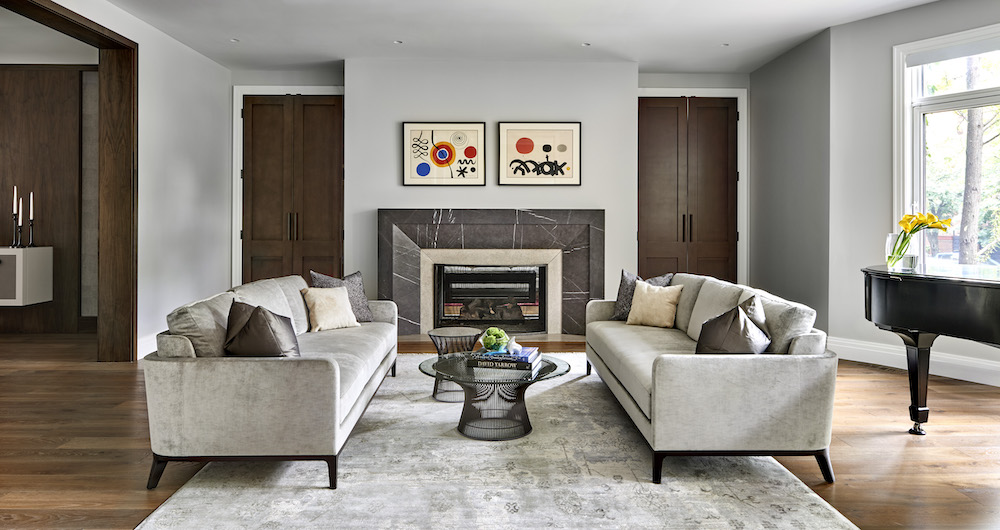 Modern living room with two grey sofas facing each other, a glass coffee table, fireplace, abstract artwork above, piano on the right, and large window letting in natural light.