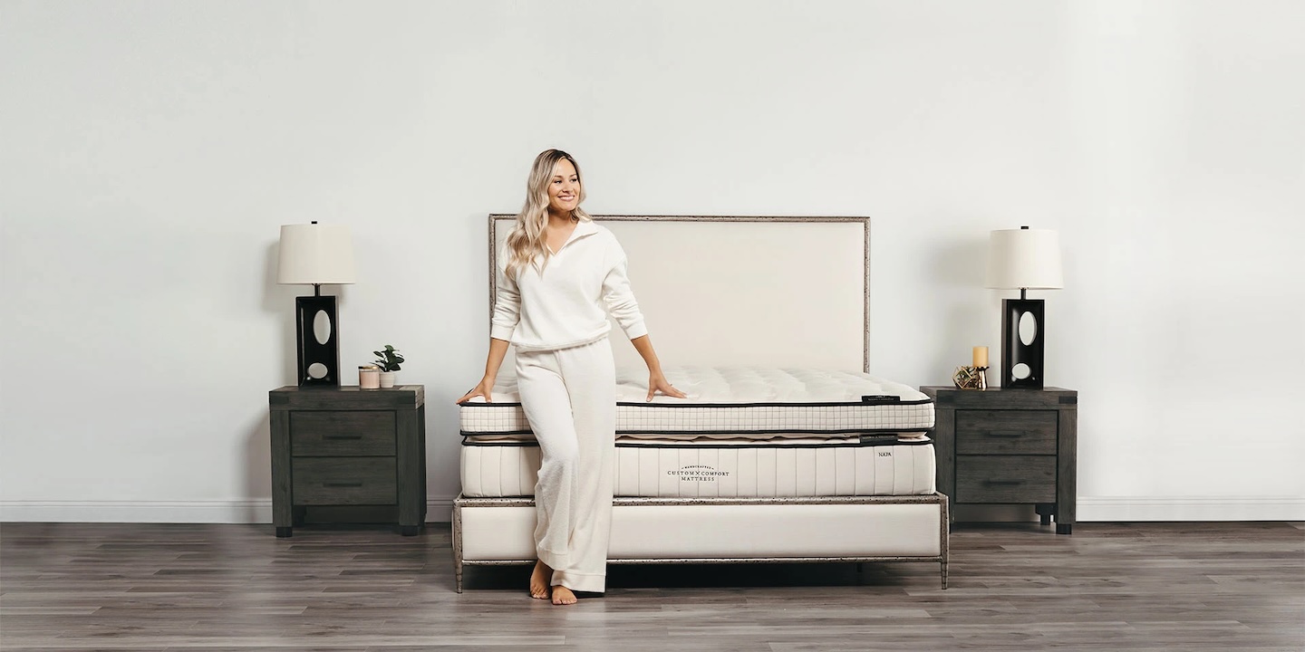A woman in white loungewear leans on a beige bed with a tall headboard. A dark nightstand with a lamp and decor sits on either side