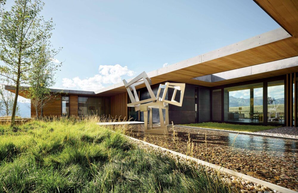 Modern house with large windows, wooden and concrete elements, grassy landscaping, and a geometric metal sculpture over a shallow water feature.