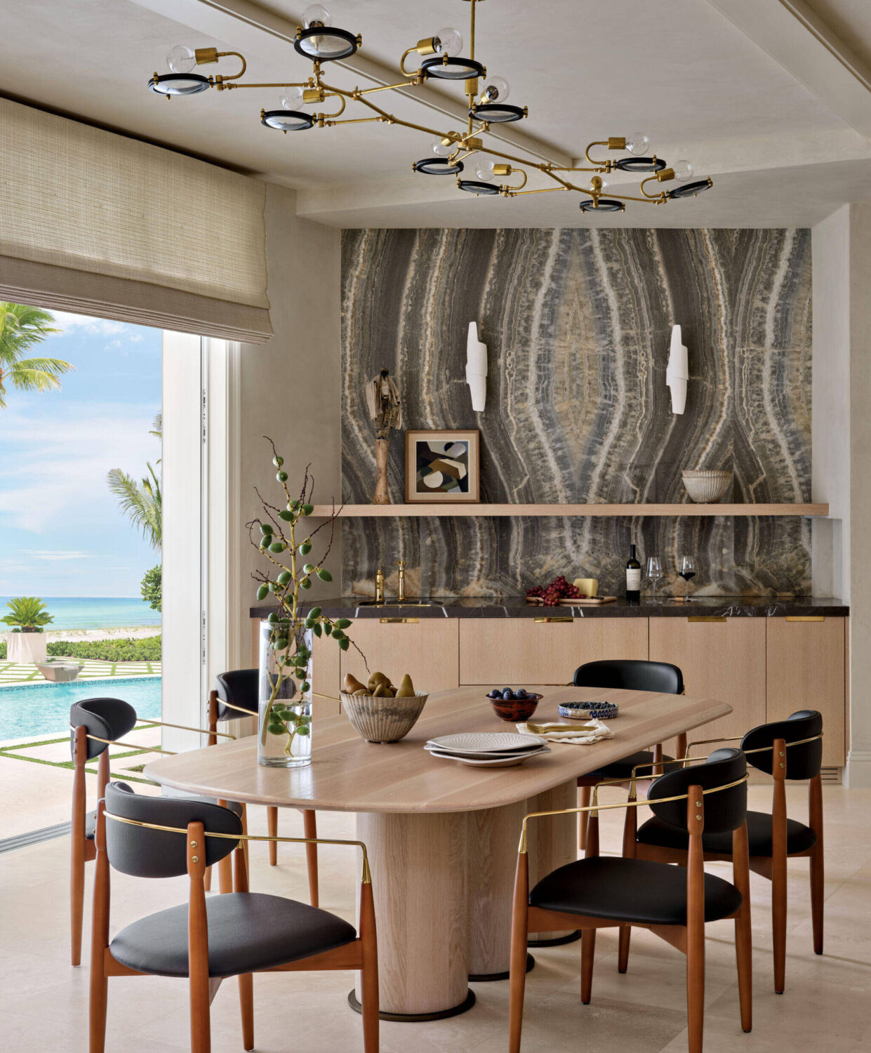 Modern dining room with a light wood table, black chairs, decorative wall, open shelving, and view of a pool and palm trees through large window.