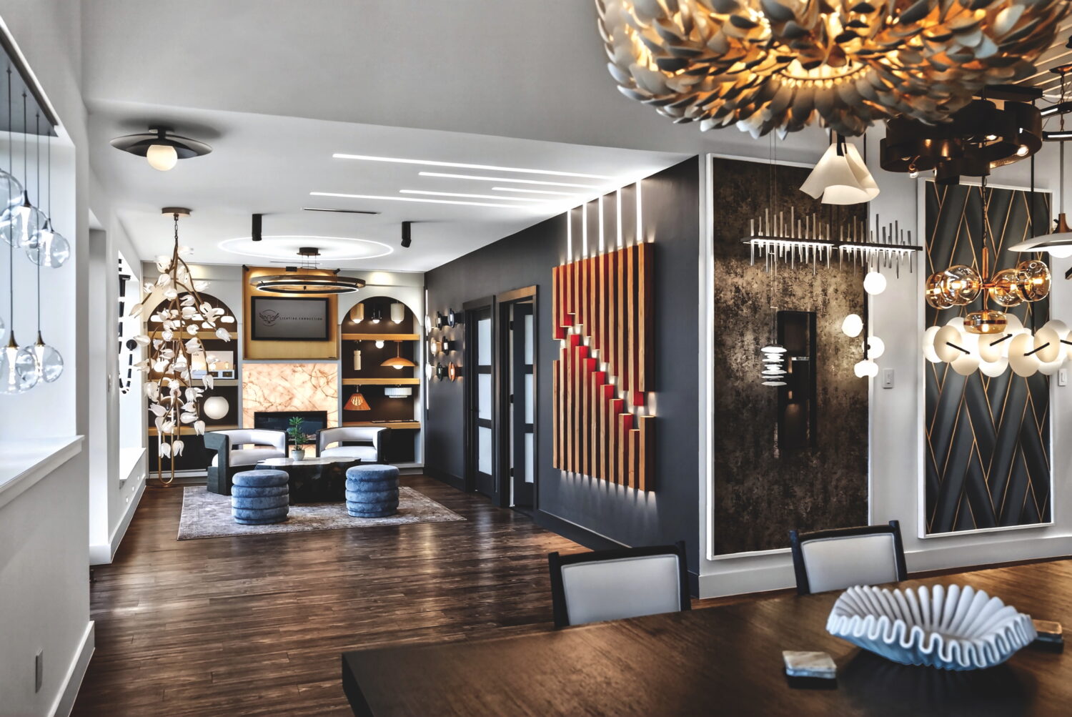 Modern interior with eclectic lighting fixtures, wall art, wood floors, and a seating area with blue ottomans and built-in shelving in the background.
