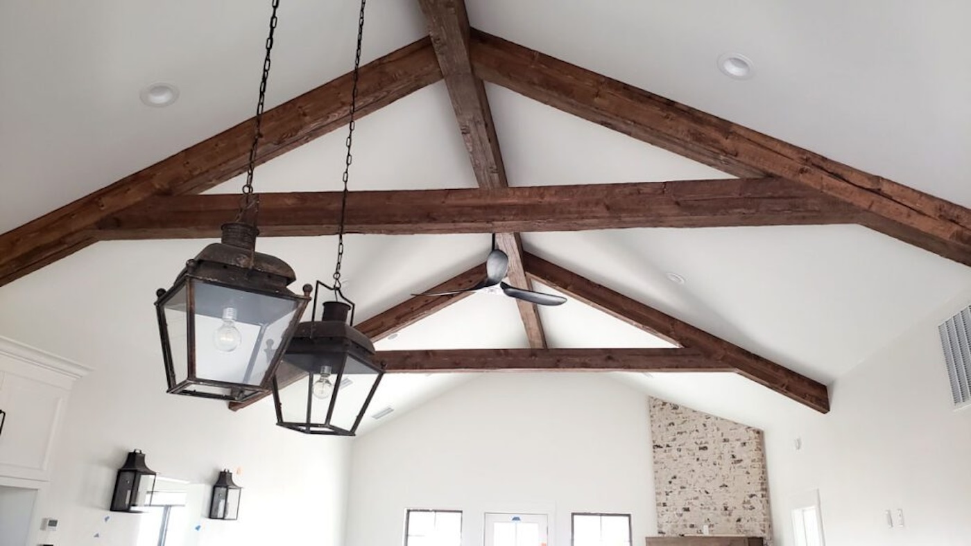 Exposed wooden ceiling beams with hanging lantern-style lights and a ceiling fan in a bright room with white walls and a brick accent wall.
