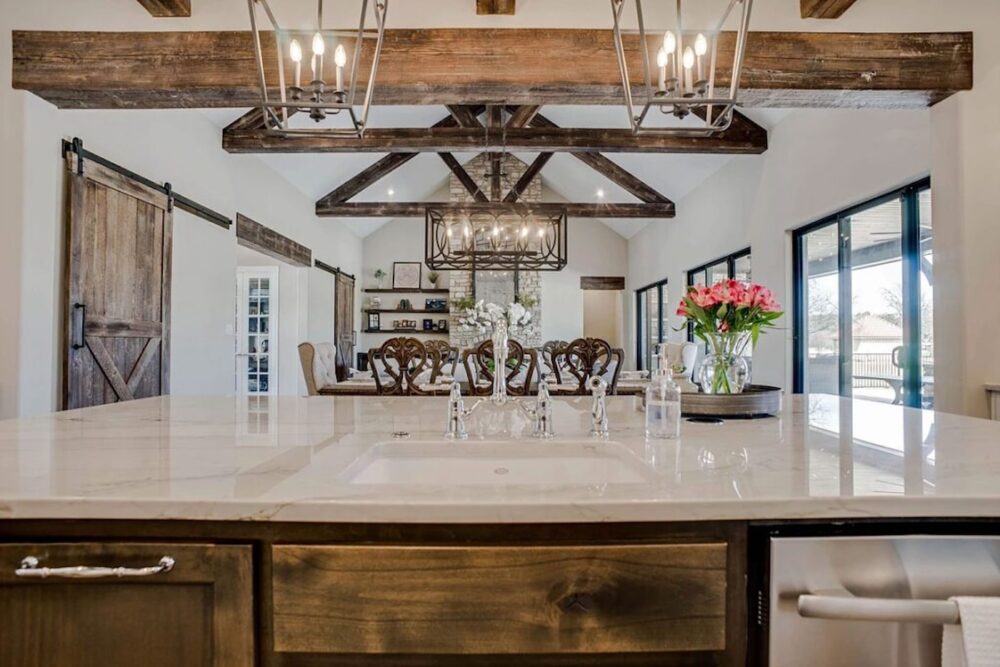 Modern farmhouse kitchen with a marble island, striking wood beams overhead, and elegant hanging lights. Pink and white flowers in vases sit on the counter, with a dining area visible in the background.