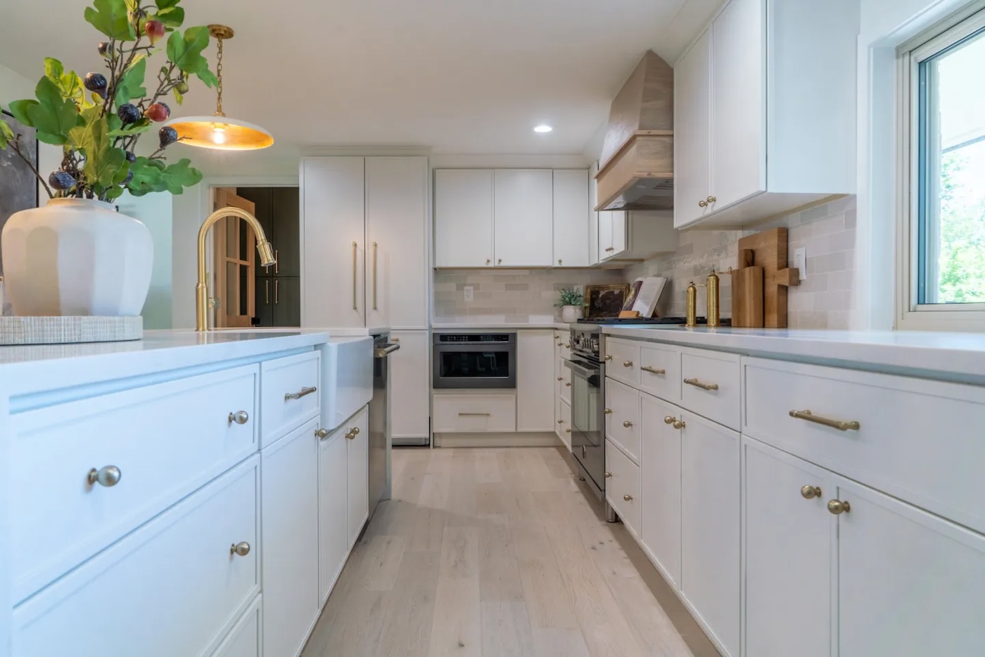 Modern kitchen with white cabinets, light wood floors, brass hardware, built-in appliances, a vase with greenery, and a window on the right side letting in natural light.
