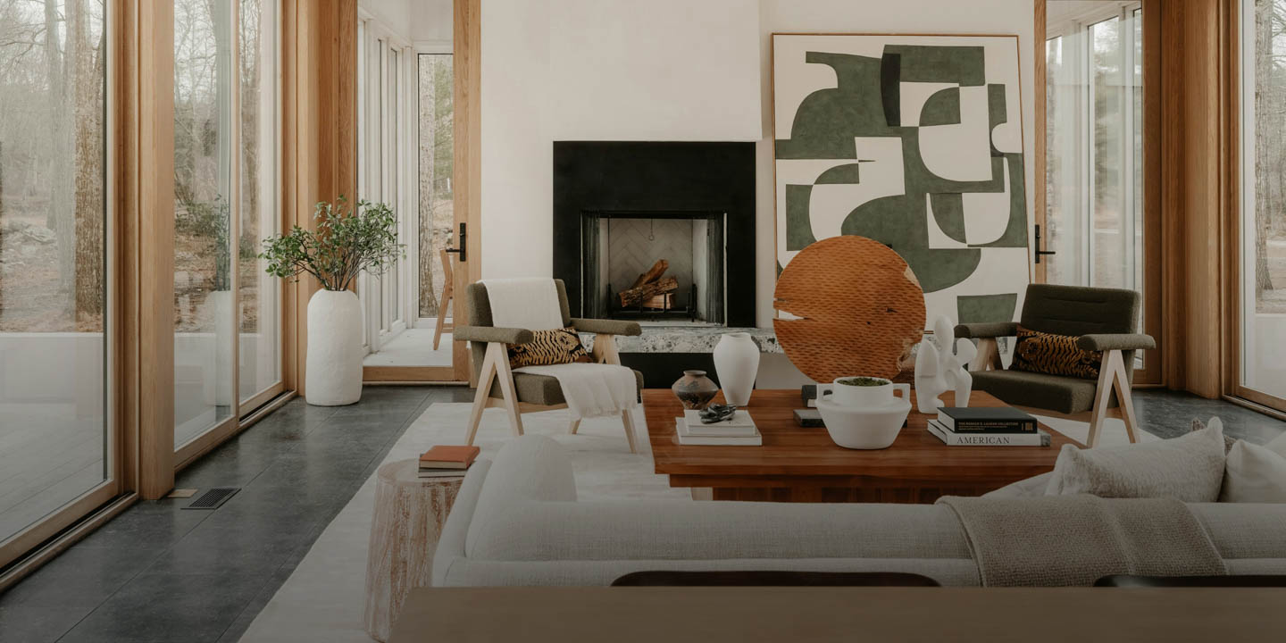 Modern living room with large windows, a central wooden table, abstract art, and a fireplace. Neutral tones create a warm and inviting atmosphere.