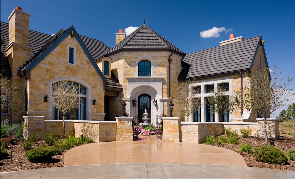 A large stone house with a dark shingled roof, arched windows, and a circular driveway under a clear blue sky.