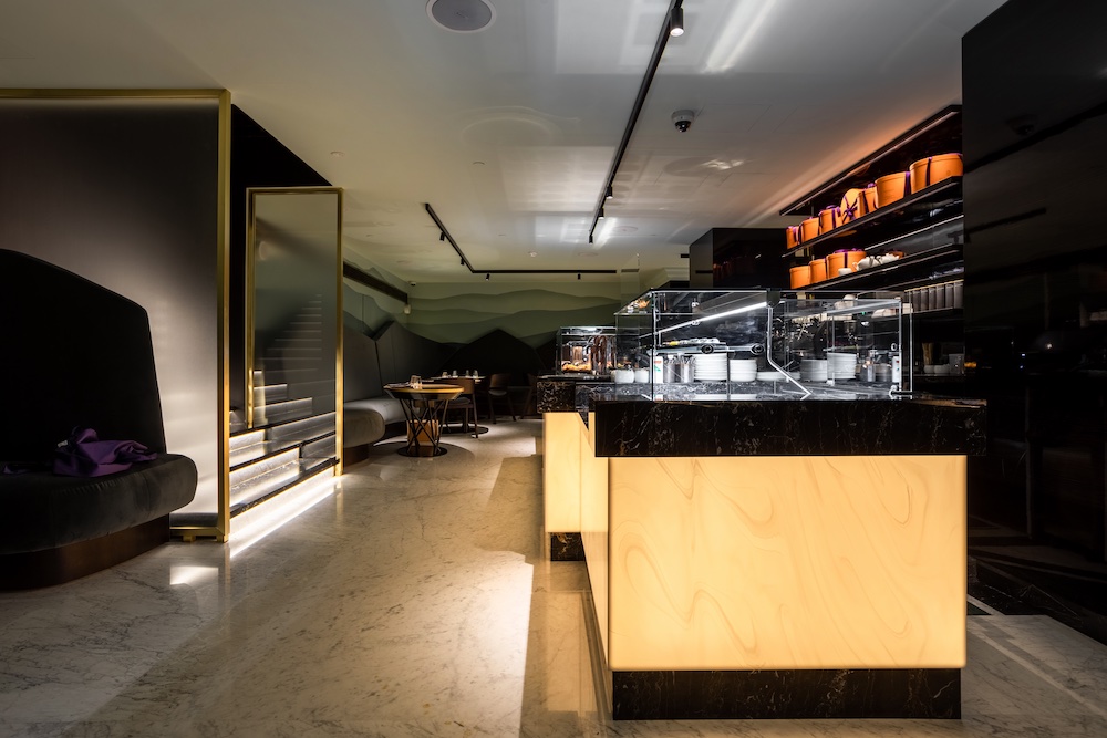 Modern cafe interior with marble floors, illuminated counter, glass pastry displays, and seating areas. Stairs and shelves with orange containers are visible in the background.
