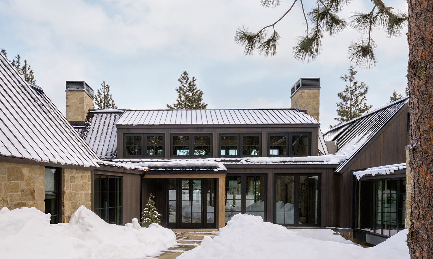 Modern house with stone and dark wood exterior, surrounded by snow.