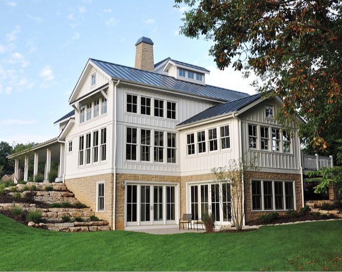Two-story modern farmhouse with white siding, large windows, and stone foundation, surrounded by grass, garden, and trees.