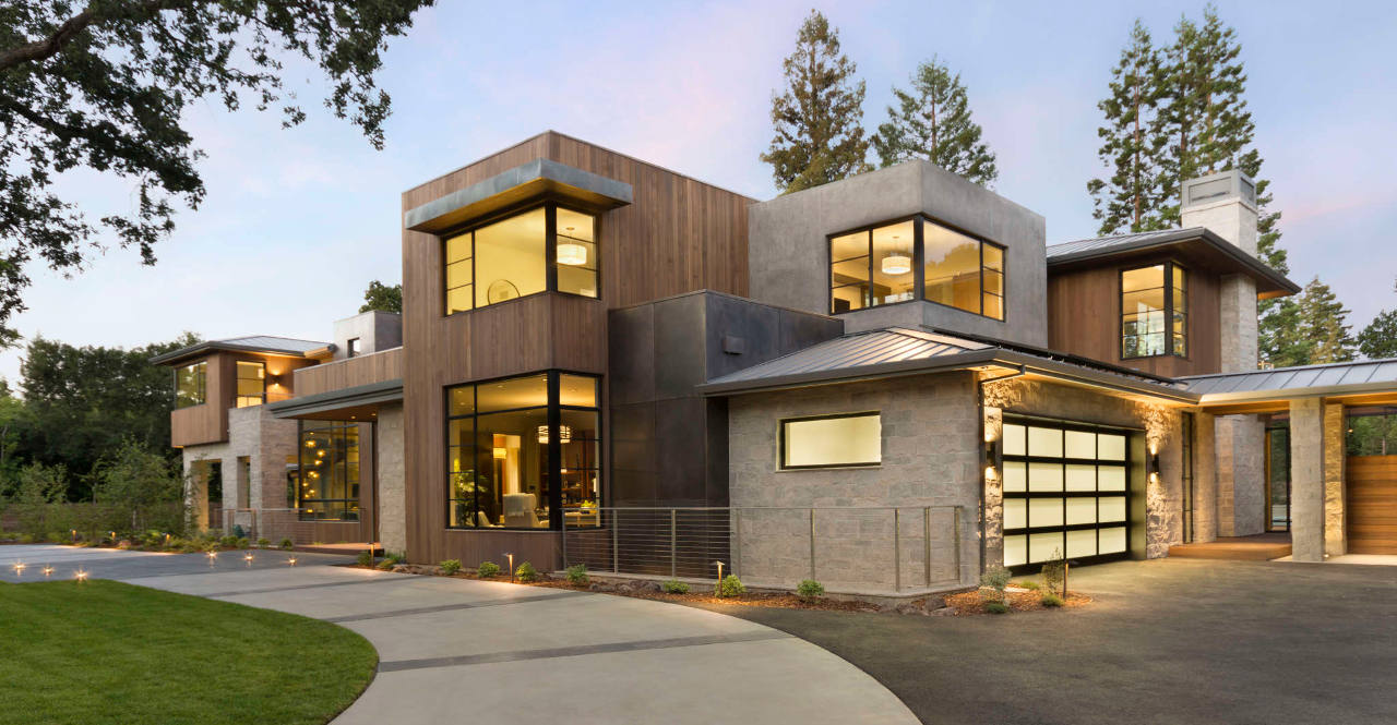 Modern two-story house with large windows, wood and stone exterior, and a driveway leading to a garage, surrounded by trees and landscaped yard.