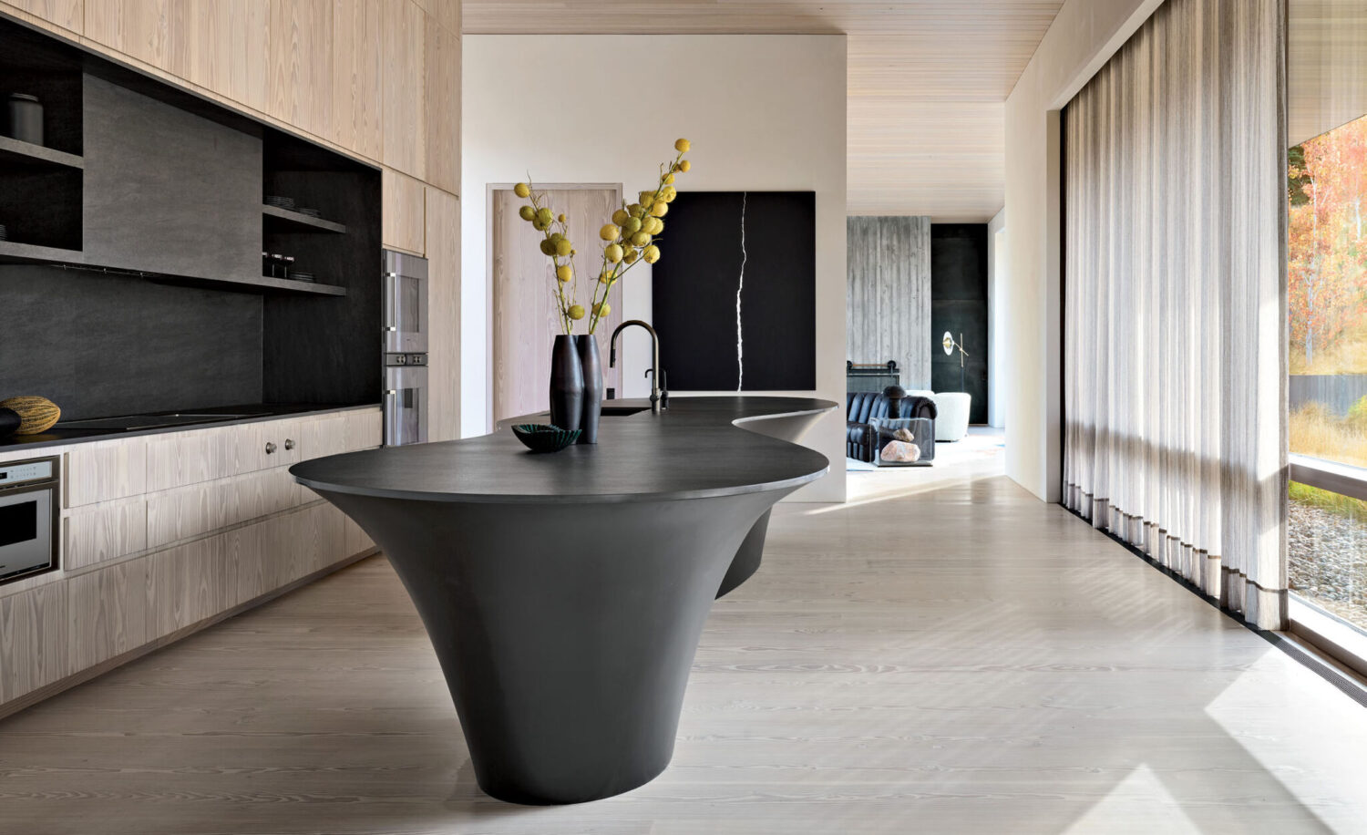 Modern kitchen with light wood cabinets, a unique black sculptural island, minimalist decor, and floor-to-ceiling windows letting in natural light.
