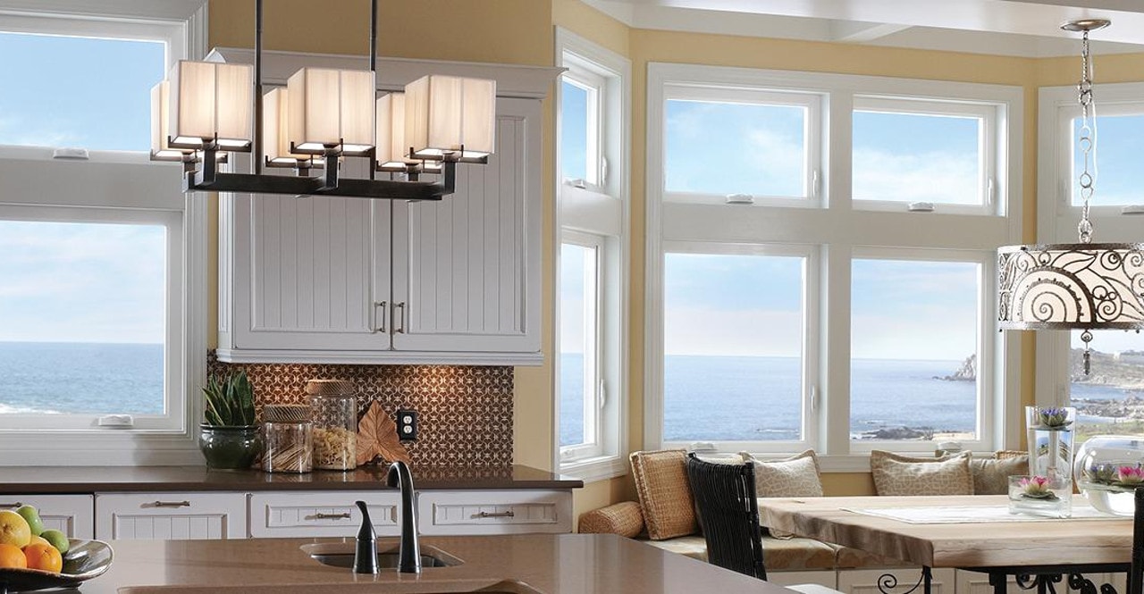 Modern kitchen with white cabinets, pendant lights, a dining table, large windows, and an ocean view in the background.