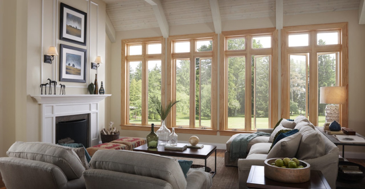 Bright living room with large windows, beige sofas, a white fireplace, artwork above the mantle, and a view of a green lawn and trees outside.